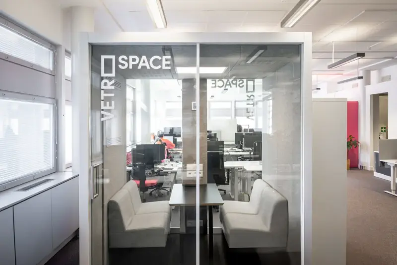 Glass-walled Vetrospace M+ pod with two sofas and a meeting table inside an open-plan office.
