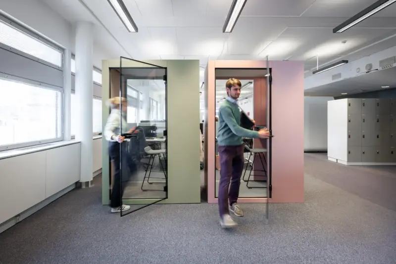 Two employees entering olive green and rose pink Vetrospace XS+ pods in a bright office.