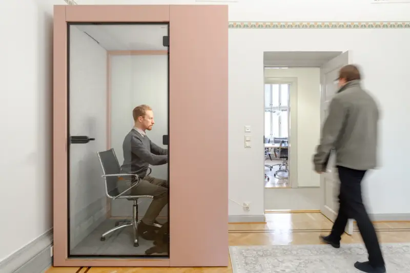 Man working inside a pink Vetrospace pod in a traditional-style office, where another man walks past.