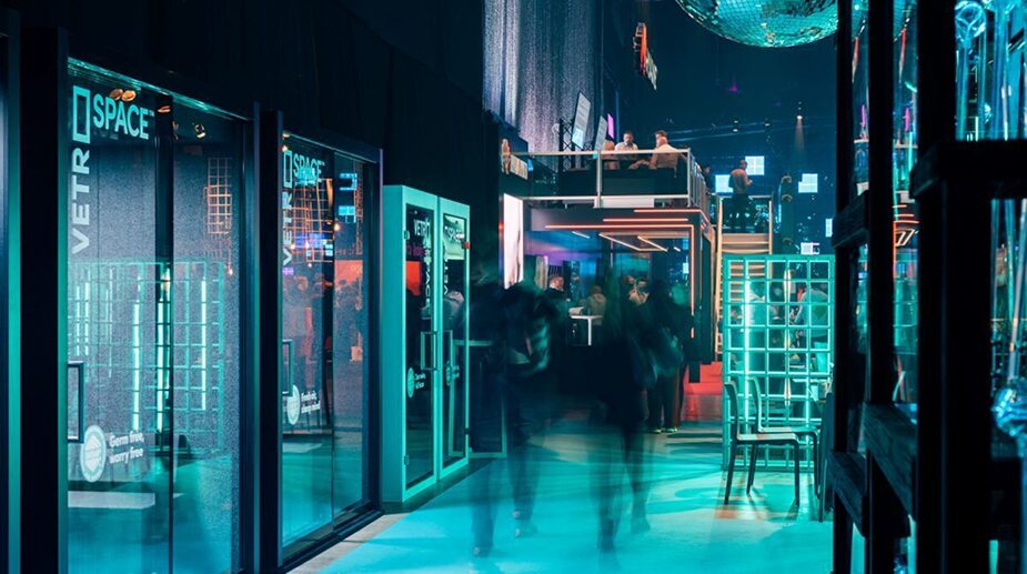 Colorfully lit event venue with people walking by modern glass-walled meeting pods, surrounded by a lively crowd and illuminated booths.