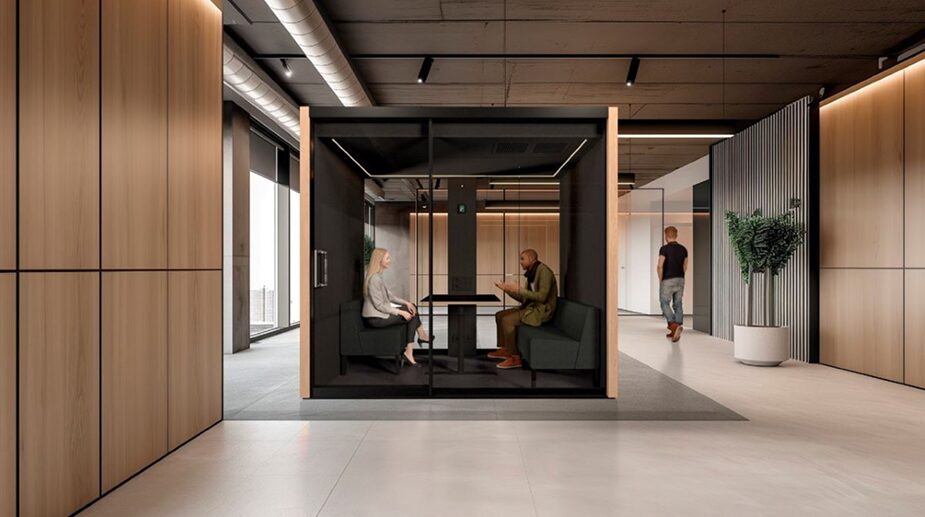 Modern office interior featuring a soundproof black-framed glass meeting pod with two professionals seated inside having a discussion, surrounded by wood paneling and minimalist decor.