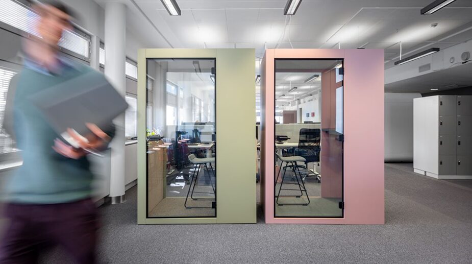Modern open-plan office space with two compact, soundproof XS+ Hybrid Pods—one green and one pink—while a blurred figure walks past holding a laptop.